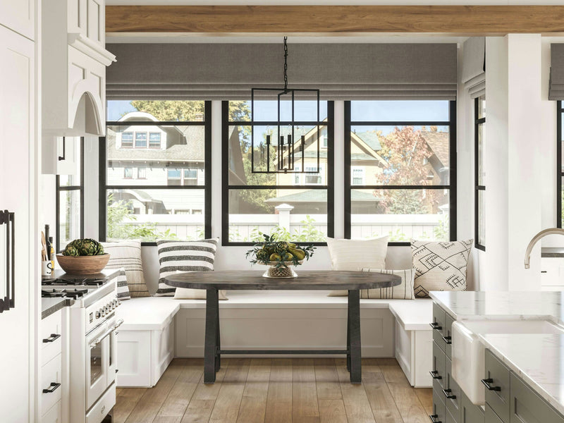 A bright kitchen with large windows and wooden floors features a cozy breakfast nook. The nook has a Wishbone Oval Dining Table in deep grey, crafted by James+James, surrounded by white built-in benches with patterned cushions. Modern appliances, cabinets, and a farmhouse-style sink complete the space.
