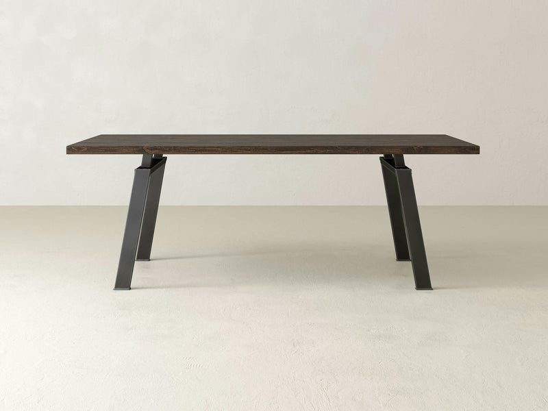 The Fulton Dining Table - Tobacco by James+James, featuring a dark brown rectangular wooden tabletop and black, angular metal legs, is placed in an empty room with light-colored walls and floor. The table boasts a sleek and modern design.