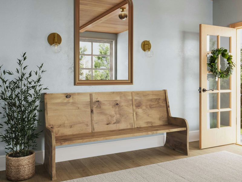 A cozy entryway featuring the Parish Pew Bench in Harvest Wheat from James+James, a large mirror, and spherical sconces on a light-colored wall. The door, adorned with a green wreath, opens to a room with abundant natural light. A potted plant sits beside the bench on a wooden floor with a light rug.