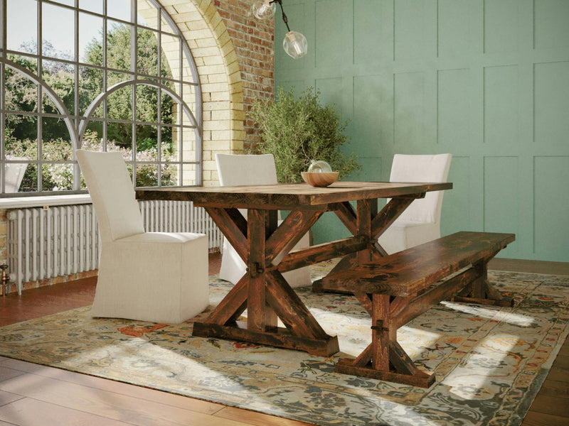 A sunlit dining room features the Trestle Dining Table - Tuscany by James+James, paired with a matching bench and two white upholstered chairs. Large arched windows reveal a lush outdoor view, and a light green paneled wall adds warmth to the space. The room is adorned with a patterned rug.