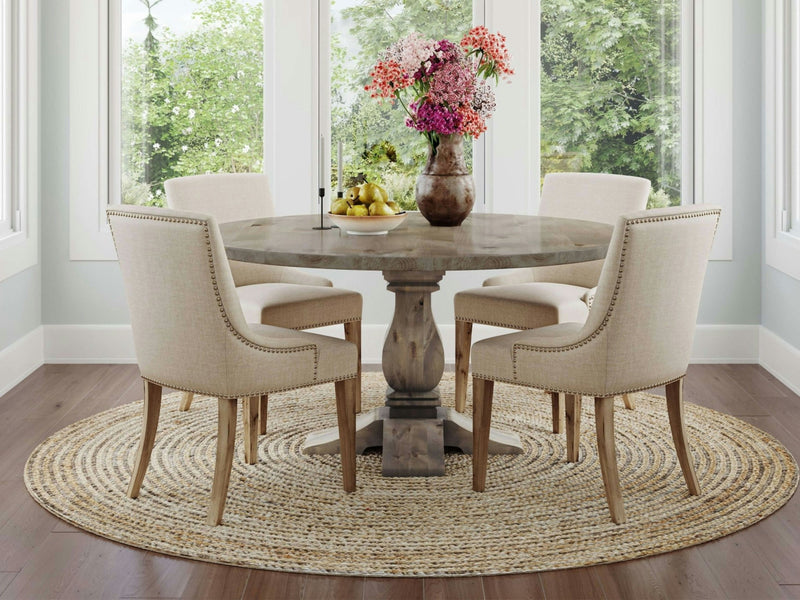 A Heirloom Round Dining Table made of Barn Wood by James+James is set in a bright room with large windows. Four upholstered chairs surround the table, which is adorned with a vase of colorful flowers and a bowl of fruit. Beneath the table lies a round woven rug, while green foliage can be seen outside the windows.