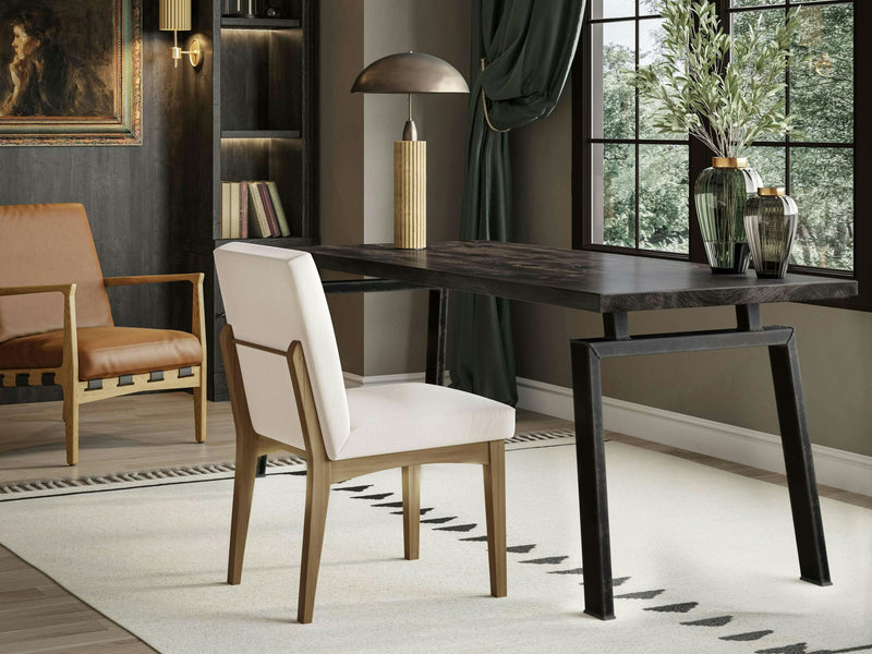 A modern home office featuring the Fulton Desk in Charred Ember by James+James, a white cushioned chair, a standing lamp, and decorative vases. A large window allows natural light to illuminate a beige rug with a geometric pattern. The space is adorned with a framed painting and a leather armchair.