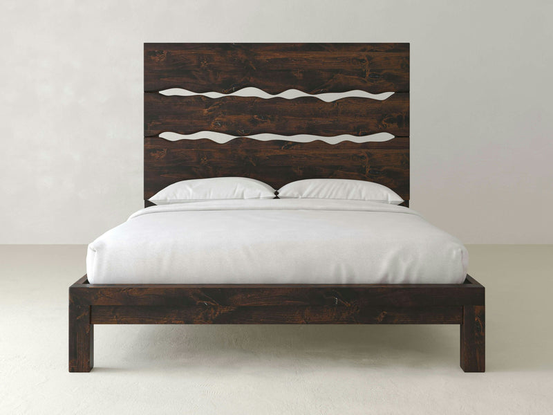 The Live Edge Bed - Tobacco from James+James features a dark, rustic finish. The headboard includes three horizontal, wavy cut-out designs. The bed is neatly made with white sheets, two white pillows, and a white comforter. The room has light-colored walls and flooring.