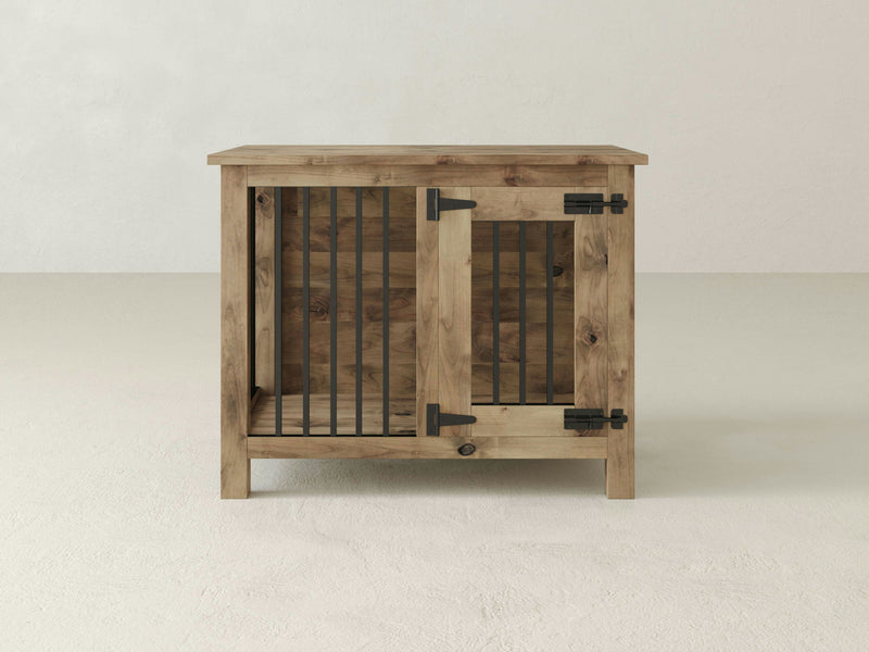 The Single Dog Kennel by James+James is crafted from Knotty Alder hardwood and showcases a rustic finish with black metal bars on the front. This wooden dog crate includes a hinged door secured with a latch, blending functionality and aesthetic appeal while resembling a small piece of furniture against a plain, light-colored wall.