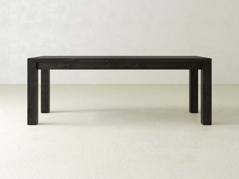 The Jonathan Desk - Charred Ember by James+James is a simple, rectangular, dark wood desk with a flat top and four straight legs. The desk is plain and unadorned, showcasing a minimalist design suitable for various home or office settings.