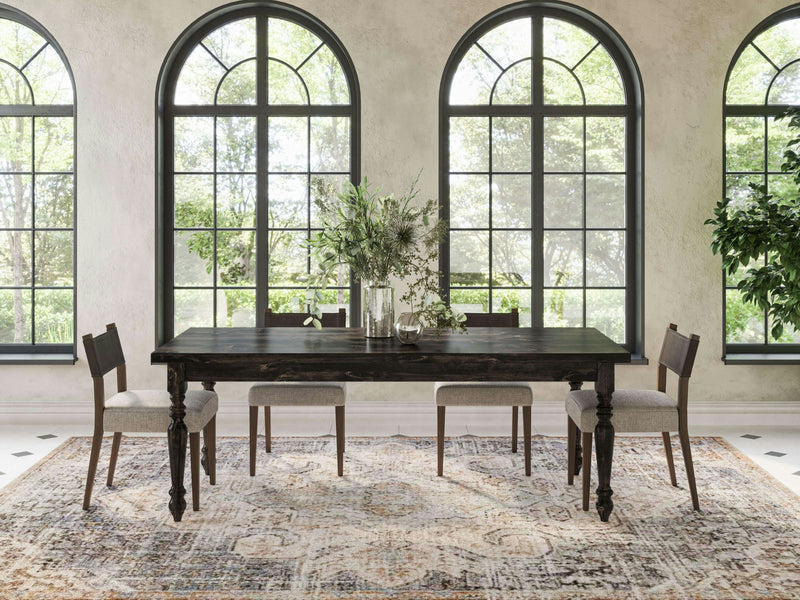 A dining room with the James+James French Country Dining Table in Charred Ember, accompanied by six chairs adorned with light-colored upholstery. The table is elegantly decorated with vases of green foliage. Large arched windows in the background let in ample natural light and offer a view of greenery outside. The floor features a patterned rug.