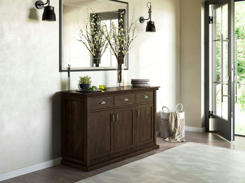 A modern, well-lit room features the Nora Buffet - Tobacco from James+James placed against a light-colored wall. Above the buffet is a large mirror. On the buffet sit various decorative items and a vase with branches. An open door to the right allows sunlight to stream in, offering a view of greenery outside.