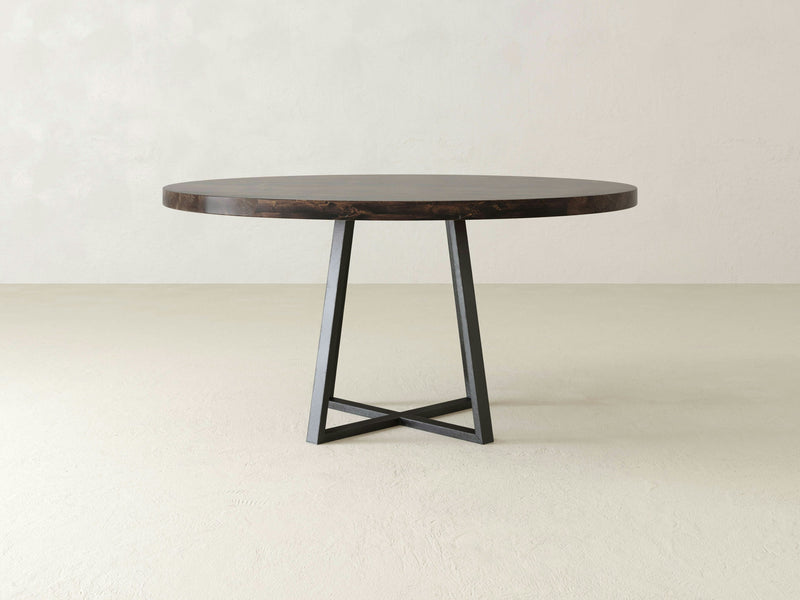 The Watson Round Dining Table - Tobacco by James+James features a dark wooden top and black metal legs arranged in a geometric, crisscross pattern, set against a plain, light-colored background. Its minimalist design lends it a modern appearance.