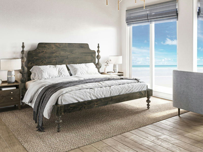 A modern bedroom with an Opal Bed in deep grey by James+James, adorned with white bedding and a gray throw blanket. The room features a light gray armchair, bedside tables with lamps, and large windows overlooking a beach with clear blue skies and ocean waves. The wooden floor is complemented by a textured rug.