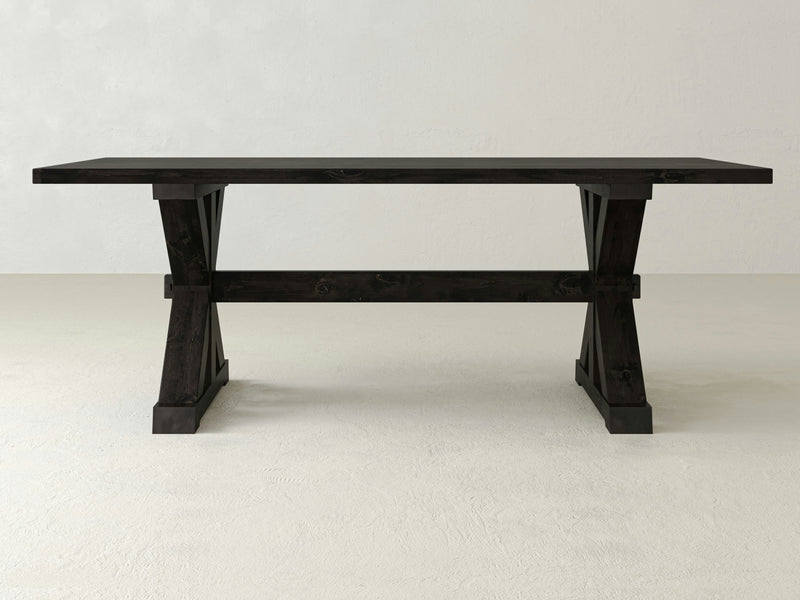 The Trestle Dining Table - Charred Ember by James+James features a dark wooden surface with a rectangular top set on trestle-style legs. This table showcases a clean, minimalist design and is placed against a plain, light-colored background.