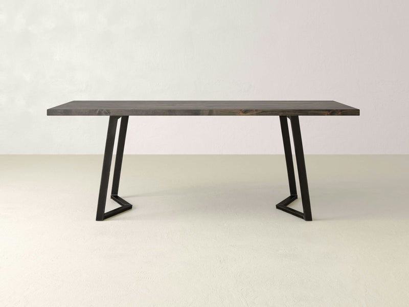 The Arkwright Dining Table by James+James is a minimalist, rectangular wooden table with a dark finish, supported by two black, angular metal legs that form a geometric design. This handcrafted dining table is showcased in a simple, light-colored modern room with no other furniture or decor visible.