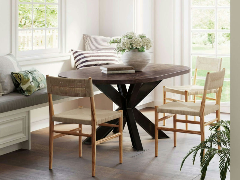 A sunlit dining area features the Shiloh Oval Dining Table - Tobacco by James+James, with its sleek modern black metal base. One side is surrounded by a cushioned bench adorned with patterned pillows, while two wooden chairs with woven seats are placed on the other side. A large vase of white flowers sits elegantly on the table.