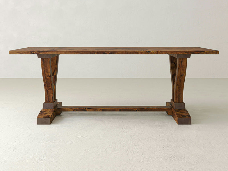 Introducing the Vera Dining Table by James+James: This exquisite dining table boasts a rectangular top and sturdy, intricately carved legs, meticulously crafted by skilled artisans. Featuring a dark brown finish and a single stretcher connecting its legs at the base, the trestle design stands out against a plain, off-white background that emphasizes its impeccable craftsmanship.