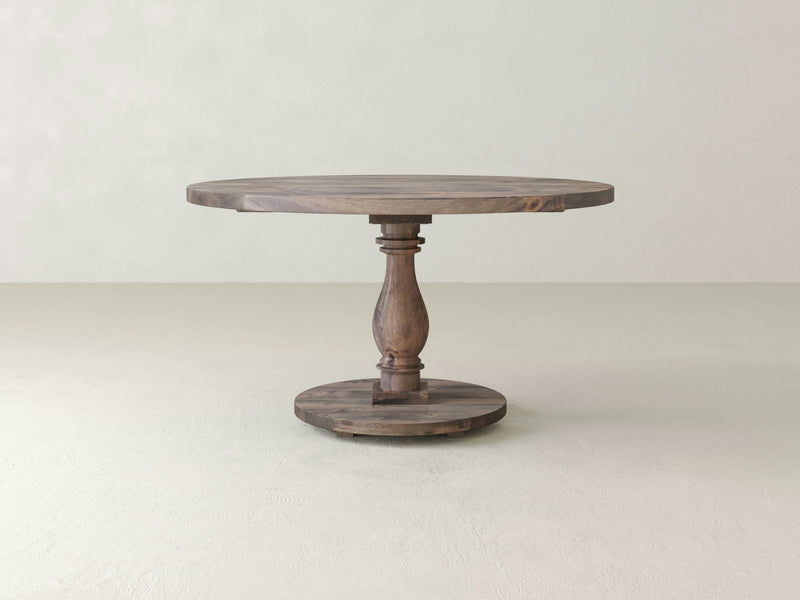 The Vivien Round Dining Table - Barn Wood by James+James is featured against a plain, light-colored background. This wooden dining table showcases a carved central pedestal base and boasts a rustic, natural finish with a simple yet elegant design.