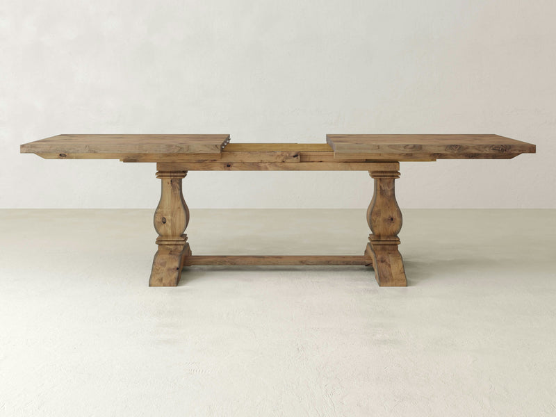 The Heirloom Expandable Dining Table - Harvest Wheat by James+James is displayed against a plain white background. This wooden dining table features a rustic finish and ornate pedestal legs, and includes an extension leaf in the center to expand its length.