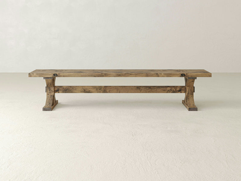 The Trestle Bench - Harvest Wheat by James+James, featuring a rustic wooden design with a simple, sturdy aesthetic, is placed in front of a plain, off-white wall. The bench has a rectangular seat and two robust legs connected by a horizontal support beam. The wood's natural finish highlights its beautiful grain pattern.