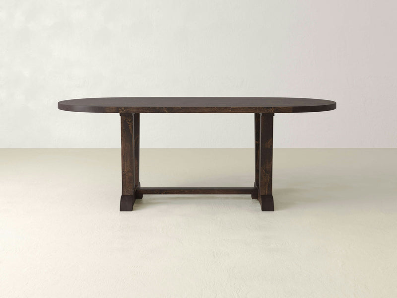 The Thaden Oval Dining Table - Tobacco from James+James features a dark brown wooden finish with an oval top and rounded edges. The table is supported by two sturdy legs connected by a horizontal stretcher, set against a plain, light-colored backdrop.