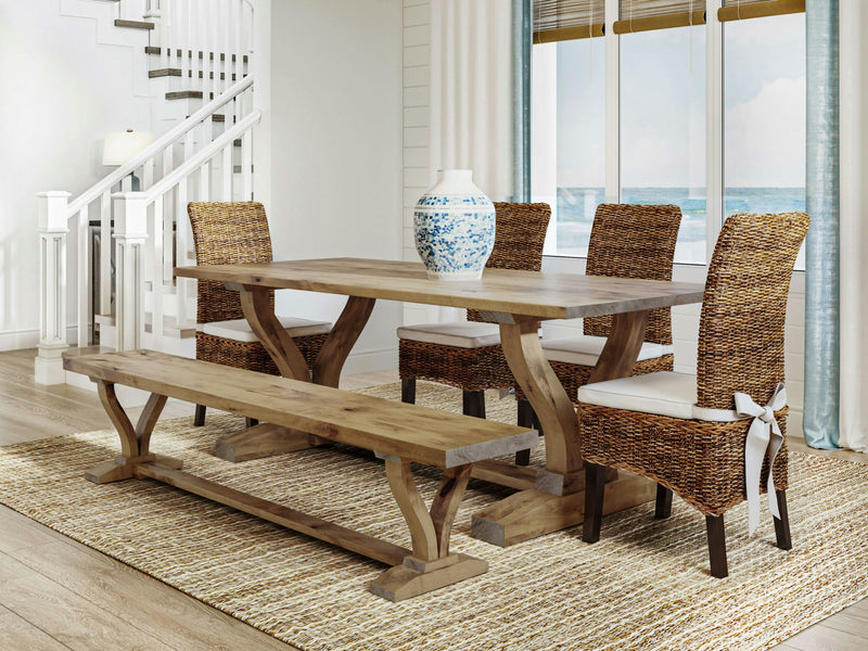 A coastal-themed dining room featuring the Vera Expandable Dining Table in Harvest Wheat by James+James, paired with a matching bench and four wicker chairs adorned with white cushions. A blue and white vase is elegantly placed on the table. The room is accentuated by a patterned rug, light-colored walls, and a window offering an ocean view.