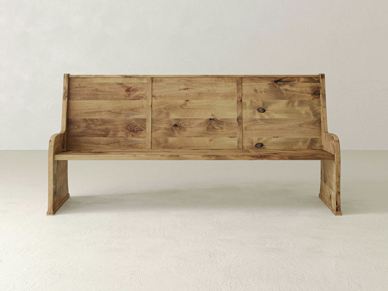 The Parish Pew Bench by James+James, featuring a vintage-style minimalist design, sits on a light-colored floor against a plain light gray wall. Resembling an antique church pew, this bench has a high backrest with three wooden panels and smooth edges on the sides, showcasing its natural wood grain and knots.