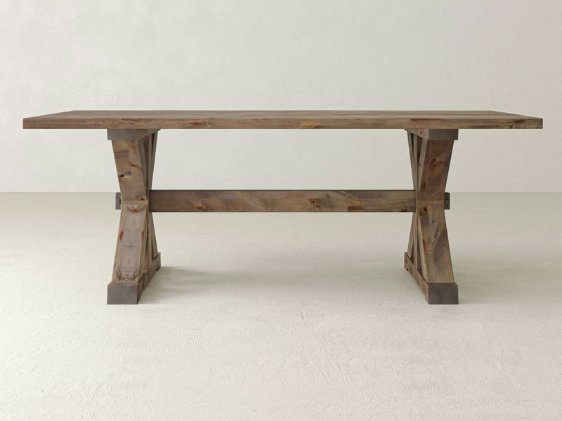 The Trestle Dining Table - Barn Wood by James+James features a rustic wooden design, complete with a rectangular top and sturdy trestle legs. The table’s natural, weathered finish highlights the wood's beautiful grain and knots. It is elegantly displayed on a light-colored floor in a minimalist room with a neutral wall background.