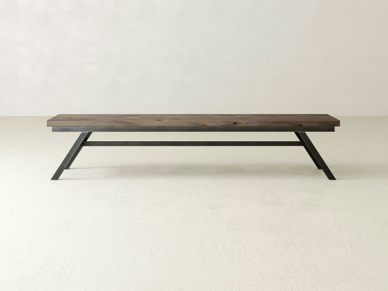 The Fulton Bench by James+James features a dark brown rectangular seat crafted from solid hardwood and black industrial steel bases that form angular supports. Placed against a plain light-colored wall on a smooth floor, it exemplifies a simple yet modern aesthetic.