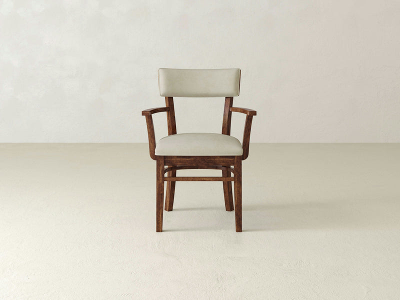 The Everly Arm Dining Chair - Tuscany by James+James, a simple wooden chair with light gray cushions, is centered in a minimalist setting. The background features a plain, light-colored wall and light floor, creating a clean and modern ambiance. The chair includes armrests and a slightly curved backrest.