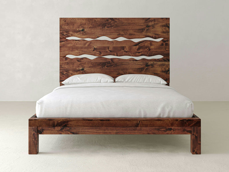 The James+James Live Edge Bed - Tuscany showcases a headboard with wave-like cut-out designs, crafted from dark wood. The bed is elegantly dressed in white bedding, complete with two pillows, set against a plain, light-colored wall.