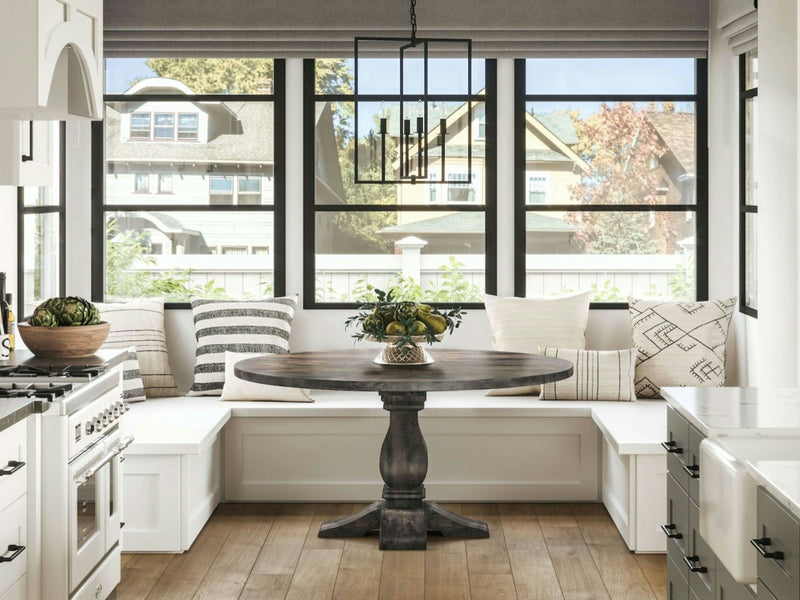 A bright kitchen nook with large windows features a Heirloom Round Dining Table in Deep Grey by James+James, adorned with a fruit bowl centerpiece and surrounded by built-in white bench seating decorated with patterned cushions. Modern light fixtures hang above, and the windows reveal a view of houses outside.