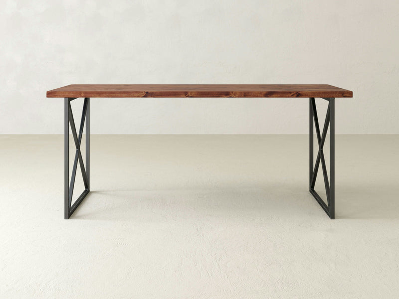 The Luca Desk - Tuscany from James+James is a minimalist wooden table featuring a dark brown rectangular top and black metal legs arranged in an X-shape. The desk is placed on a smooth, light-colored floor against a plain white wall.