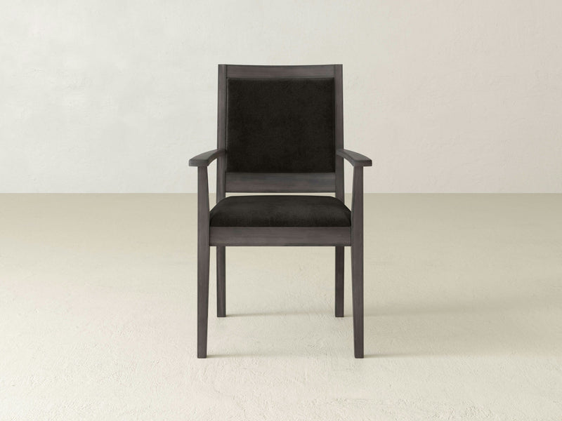 The Gina Arm Dining Chair in Deep Grey by James+James is centered against a plain off-white background. This elegant chair features a dark wooden frame with black cushioned seat and backrest, and showcases a simple, contemporary design with armrests and straight legs.