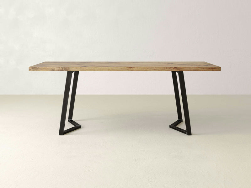 A minimalist Arkwright Dining Table - Harvest Wheat by James+James features a natural wooden finish with a rectangular top. The table stands on two black metal legs angled and connected at the base, forming geometric shapes. The background is a plain off-white wall with a beige floor.