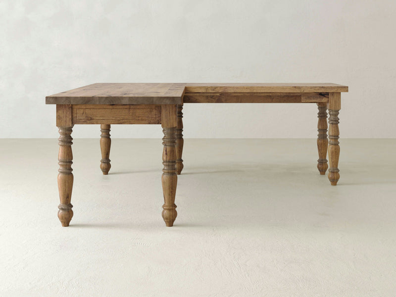 A Turned Leg Corner Desk - Harvest Wheat by James+James stands against a plain light-colored wall. The desk's surface appears smooth, showcasing a natural wood finish with visible grain patterns and intricately carved legs.