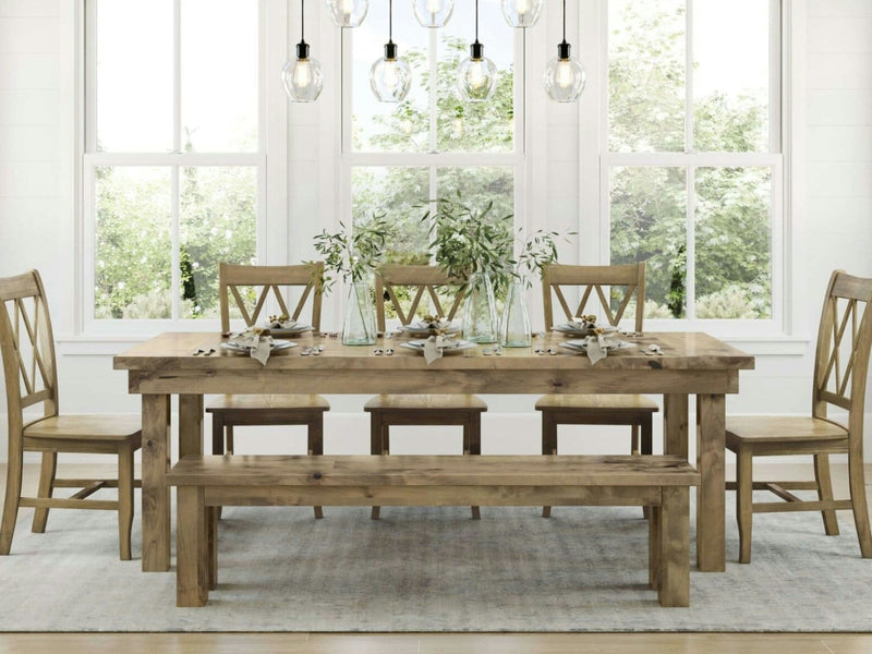 The James+James Farmhouse Dining Table in Harvest Wheat, paired with six chairs and a bench, is set with plates, cutlery, and glass jars filled with greenery. The room boasts large windows offering outdoor views, white walls, and hanging pendant lights. A light-colored rug lies beneath the table.