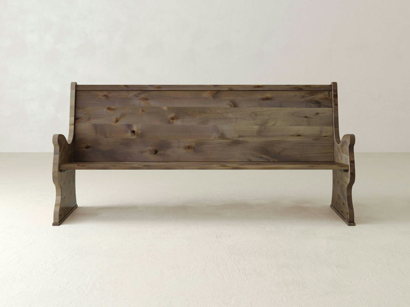 A Rustic Church Pew Bench - Barn Wood from James+James, featuring a high backrest and armrests on both sides, is placed against a plain light-colored wall on a white floor. The bench boasts a rustic appearance with a natural wood grain texture.