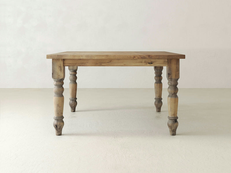 A Baluster Square Dining Table - Harvest Wheat by James+James with a rectangular top and turned legs, standing in an empty, neutral-colored room. The table features a natural, rustic finish that highlights the wood grain.