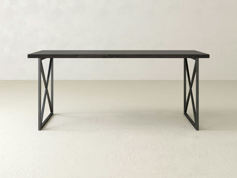 The Luca Desk - Charred Ember by James+James, a minimalist rectangular dark-colored table with a flat top and black metal legs forming an X-shape on each side, stands on a light-colored floor against a plain wall.