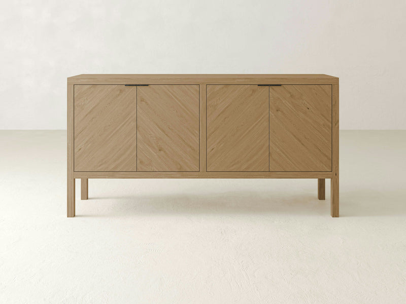 Introducing the White Oak Buffet - Barn Wood by James+James. This light wood sideboard showcases a minimalist design, featuring four doors with subtle chevron patterns and no visible hardware. It stands on four slender legs against a neutral, light-colored wall and floor, enhancing the simple and modern aesthetic.