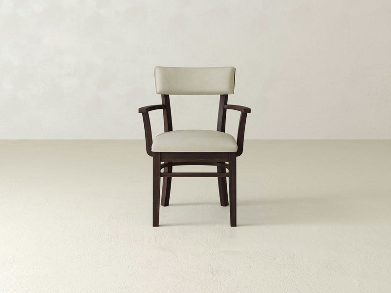 An Everly Arm Dining Chair - Tobacco by James+James, featuring a dark wooden frame and a light beige cushioned seat and backrest, is placed in a minimalist setting with a light neutral background. The chair includes armrests and boasts a sturdy design, with no other objects or decor present.