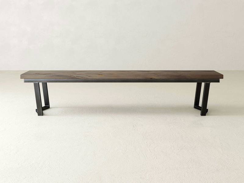 The Arkwright Bench - Barn Wood by James+James stands against a plain, light-colored background, showcasing its minimalist design. The bench features a dark brown seat made of barn wood and sleek, black metal legs. Its simple and modern aesthetic includes clean lines without armrests or a backrest.