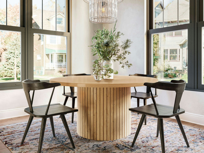 A close-up view of the Arthur Dining Table - Satin by James+James showcases its smooth, round tabletop. The base features a vertical slat design, giving it a modern and stylish appearance. The wood has a natural finish, highlighting its grain and texture against a white background.