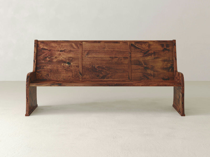 The Parish Pew Bench - Tuscany by James+James, featuring a simple and rustic design with a high backrest, stands on a smooth, light-colored floor against a plain, off-white background. The bench boasts a rich, dark brown finish with visible wood grain patterns.