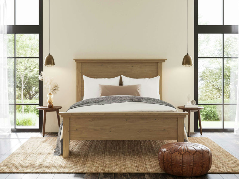 A cozy bedroom featuring the Ivy Bed - Satin by James+James, adorned with white bedding and a gray throw blanket. Flanking the bed are two bedside tables decorated with various items, each illuminated by hanging pendant lights. A brown pouf rests on a woven rug at the foot of the bed, while large windows provide an outdoor view.