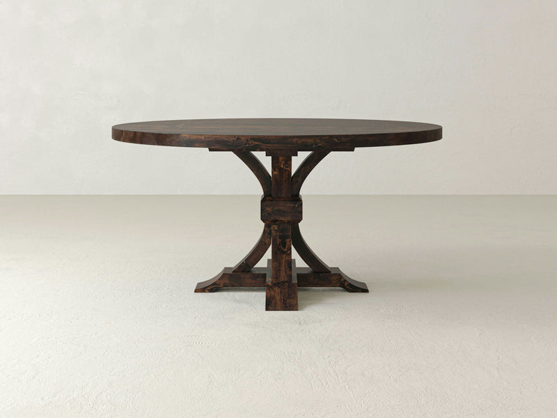 The Violet Round Dining Table - Tobacco by James+James features a round, dark wooden design with a rustic finish. This table boasts a thick, solid top and a distinctive pedestal base composed of four curved legs that connect centrally and flare out at the bottom for stability. The table is showcased on a white background.