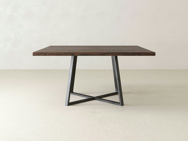 In an empty, light-colored room, the Watson Square Dining Table - Tobacco by James+James stands elegantly with its minimalist design. Featuring a dark wooden top and a geometric black metal base, this table's clean lines contribute to its contemporary appeal.