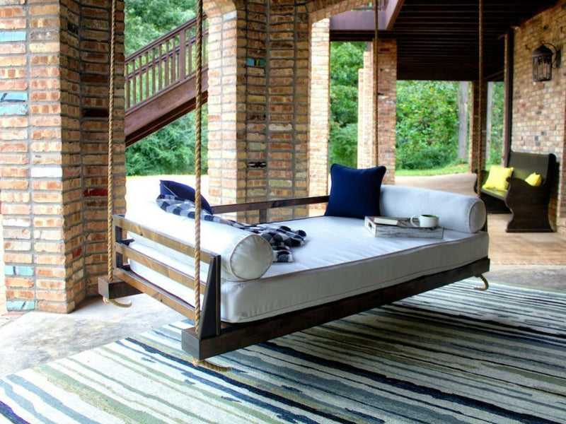 The James+James Piper Porch Swing in Tobacco, featuring white cushions and navy blue pillows, hangs securely by thick ropes under a covered brick patio. A striped rug is positioned beneath the swing, with chairs and a table seen in the background. Green foliage can be observed beyond the patio area.