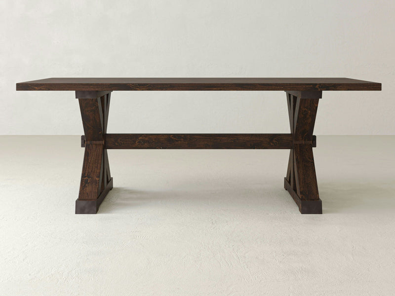 The Trestle Dining Table - Tobacco from James+James stands against a plain white background. This wooden table features a rectangular top, sturdy legs with a trestle base, and cross braces for added support. The design is both minimalist and rustic with its dark brown finish.