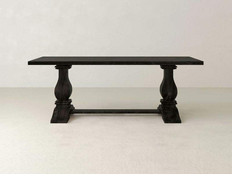 The Heirloom Dining Table - Charred Ember by James+James features a large, rectangular wooden top with a dark finish. It is supported by two thick, intricately carved pedestal legs connected by a horizontal stretcher. The table is set against a plain, white wall with a light-colored floor.