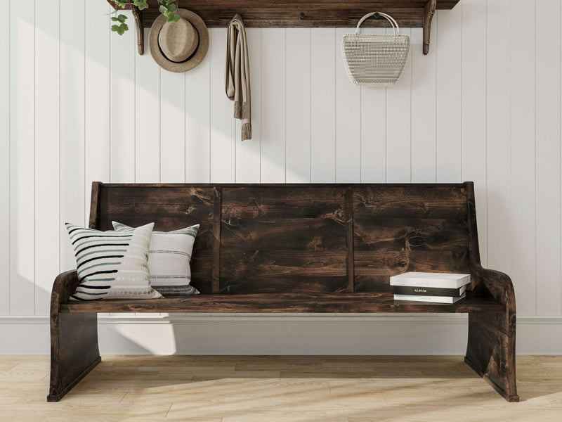 A rustic Parish Pew Bench in Tobacco finish from James+James sits against a white paneled wall, adorned with a striped pillow and a textured cream pillow. Above, a wooden shelf holds a hanging hat, a small plant, and a woven handbag. A box labeled "ALOHA" sits on the bench's right side.