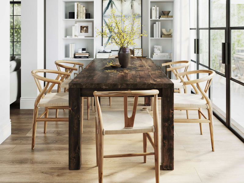 A dining room featuring the Jonathan Dining Table - Tobacco by James+James, surrounded by six light wooden chairs. A dark vase filled with yellow flowers sits at the center of the table. In the background, white bookshelves adorned with various decorations add to the charm. Large windows on the right allow natural light to flood in.