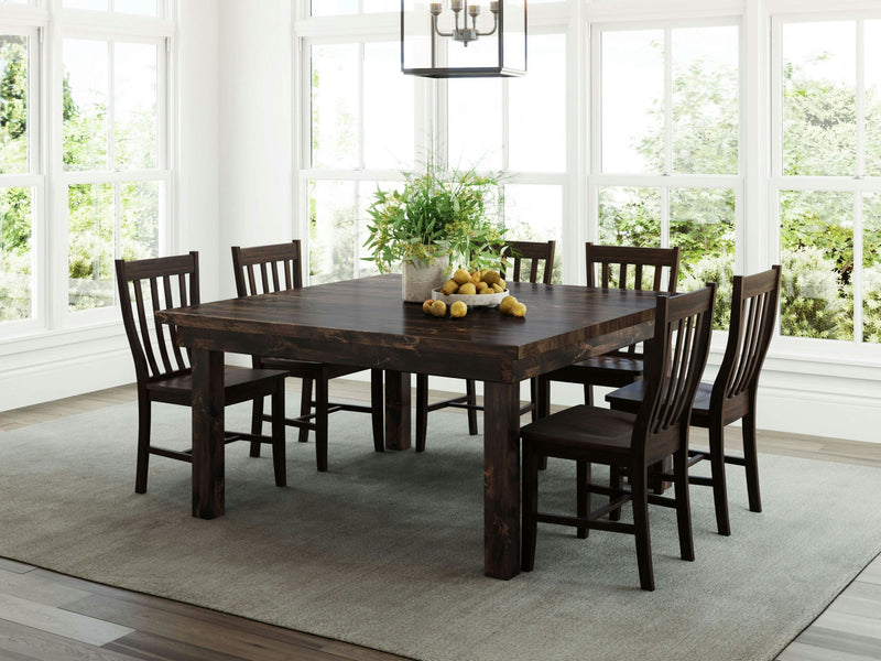 A spacious dining room featuring the Farmhouse Square Dining Table in Tobacco by James+James, surrounded by six matching chairs. The table is adorned with a potted plant and a bowl of lemons as a centerpiece. The room is bright with natural light streaming through large windows, and the floor is covered with a light-colored area rug.